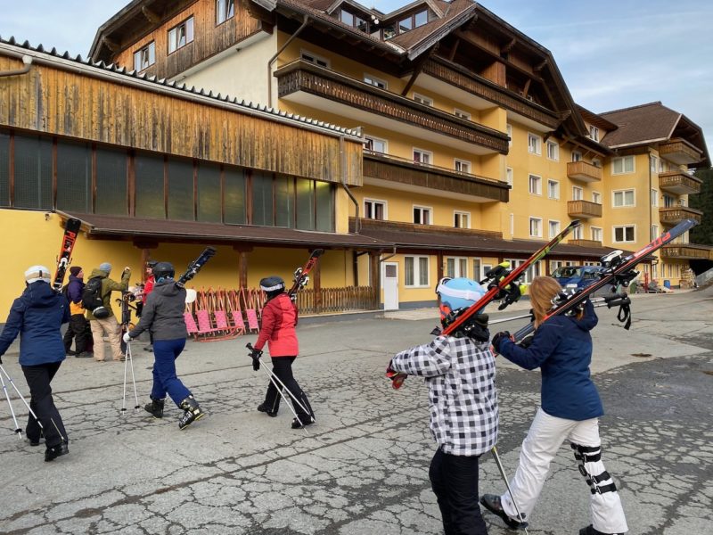 A group of men, women, children crossing a road wearing ski clothes and carrying skis on shoulders