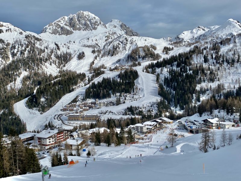 Wide shot, buildings in a snowy landscape showing ski runs with tiny figures skiing, cloudy sky, long shadowns on the ski run 