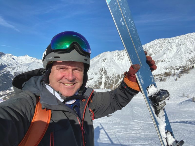 James, wearing helmet, goggles and dark grey jacket is grinning widely as he holds up a pair of blue wedze skis